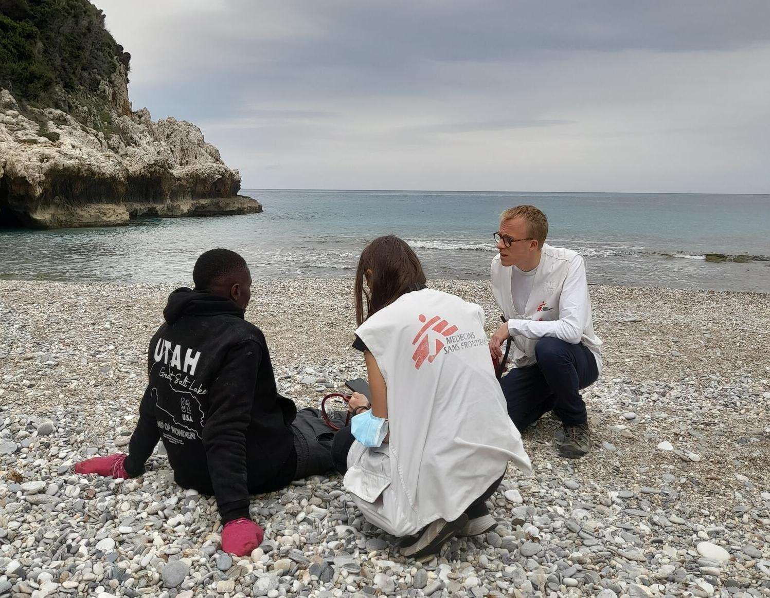 A man sits on the shores of Greece and is tended to by two MSF workers.
